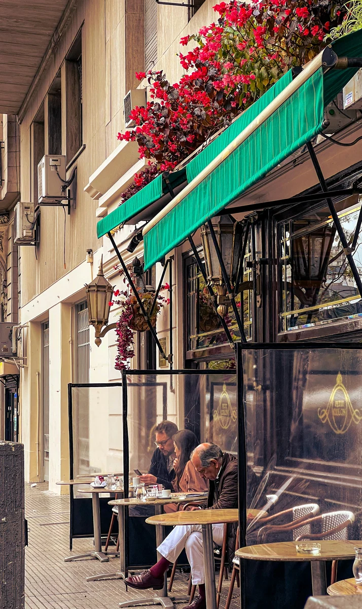 Gäste an kleinen Straßencafé-Tischen unter einer grünen Markise mit roten Blumen in Buenos Aires.