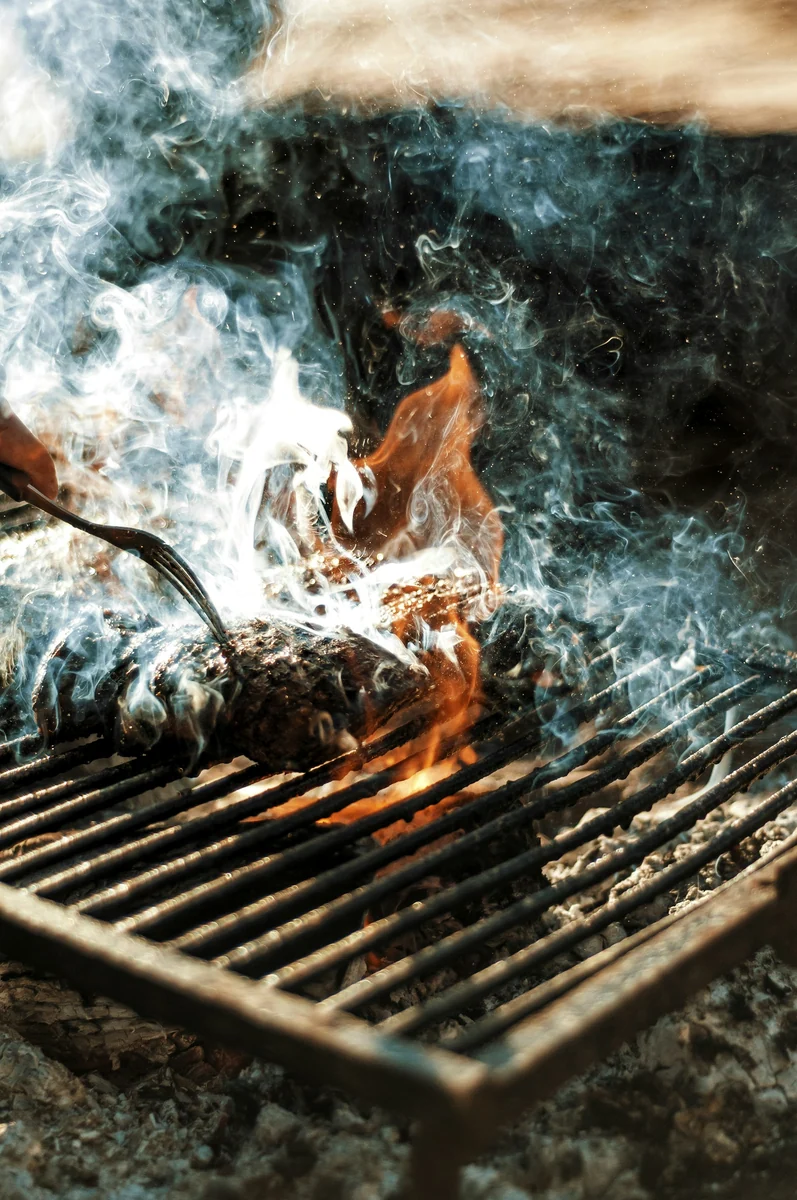 Fleisch auf einem offenen Grill, darüber Rauch und Flammen.