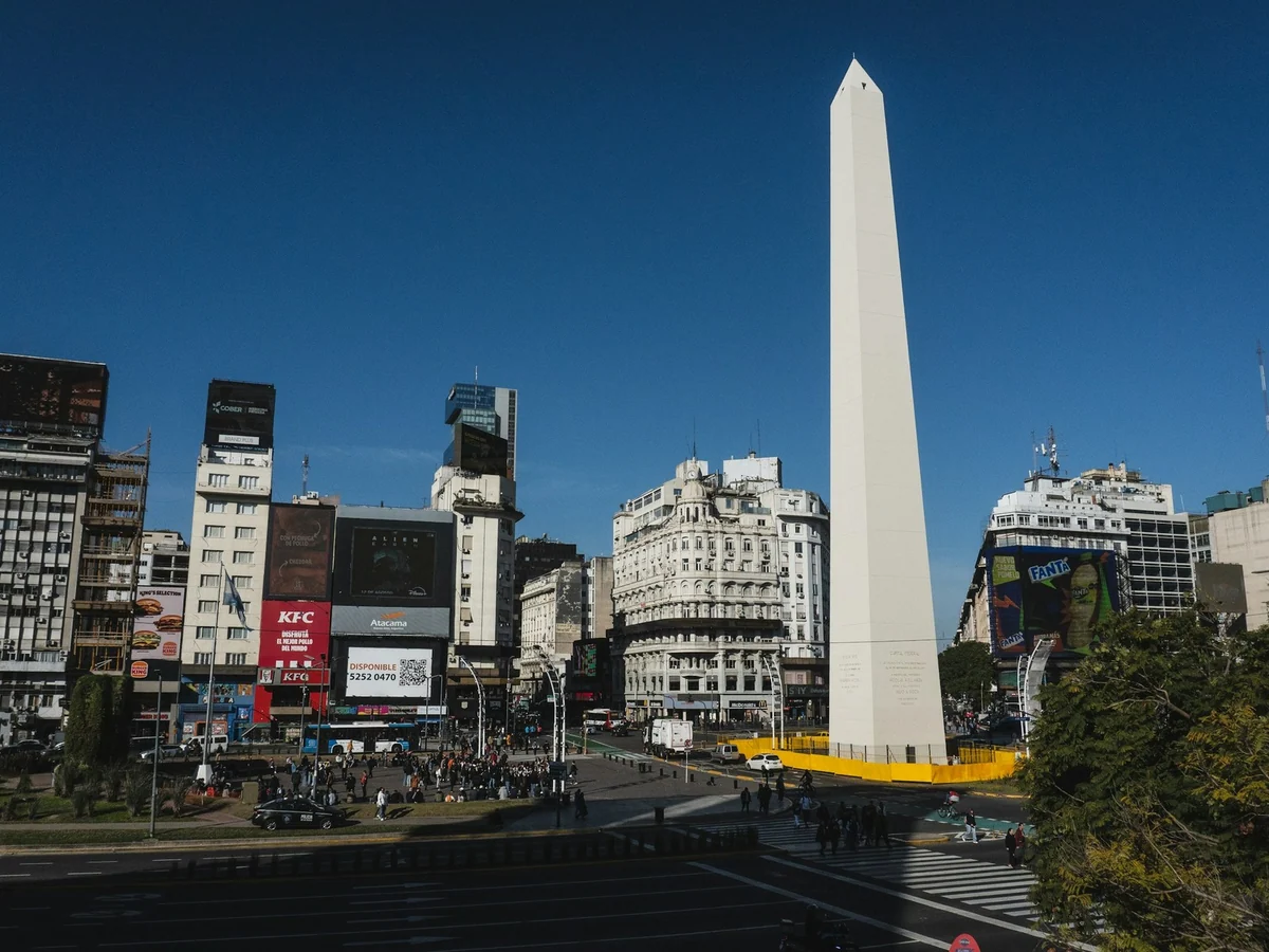 Der Obelisco vor den Fassaden und dem Straßenverkehr im Zentrum von Buenos Aires unter blauem Himmel.