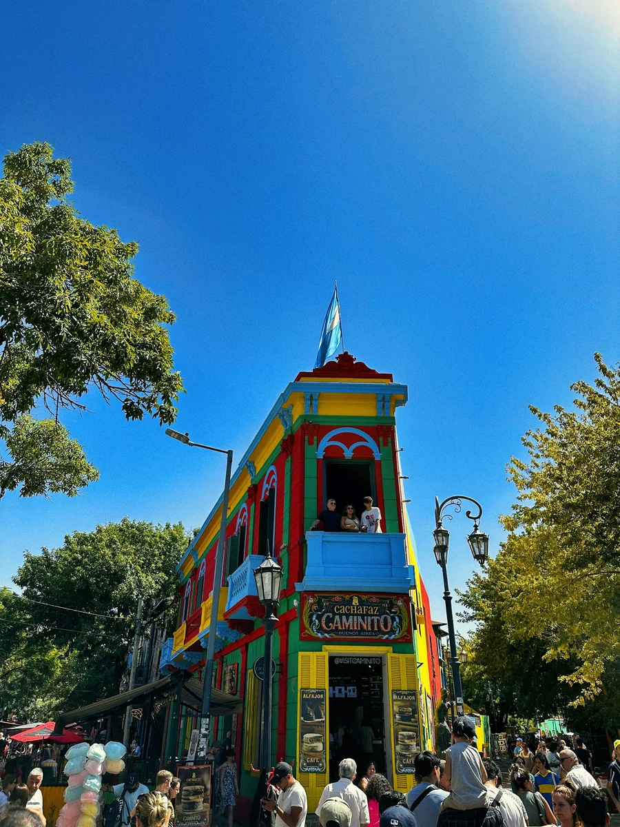 Bunt gestrichenes Eckhaus am Caminito in La Boca unter blauem Himmel, davor Besucher.