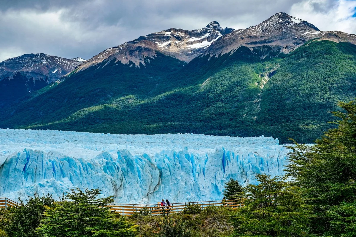 Blaue Eisfront des Perito-Moreno-Gletschers vor bewaldeten Bergen, im Vordergrund die Besucherstege.