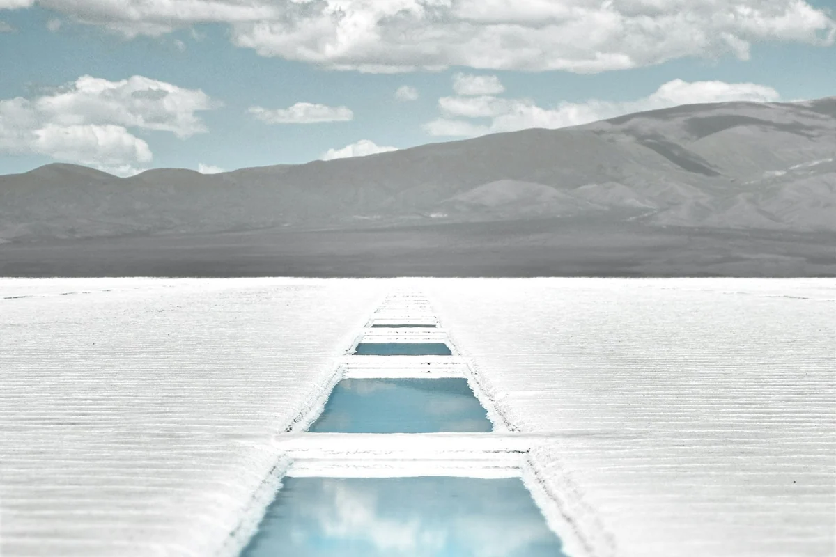 Türkisfarbene Salzbecken ziehen sich durch die weiße Ebene der Salinas Grandes vor kahlen Andenbergen.